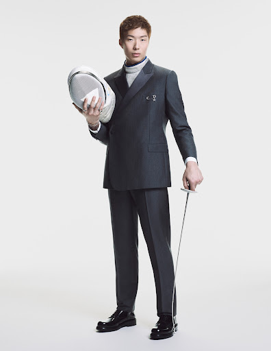 Asian man in a grey double breasted suit holding a fencing mask and foil against a white background