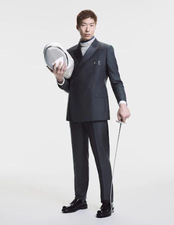 Asian man in a grey double breasted suit holding a fencing mask and foil against a white background