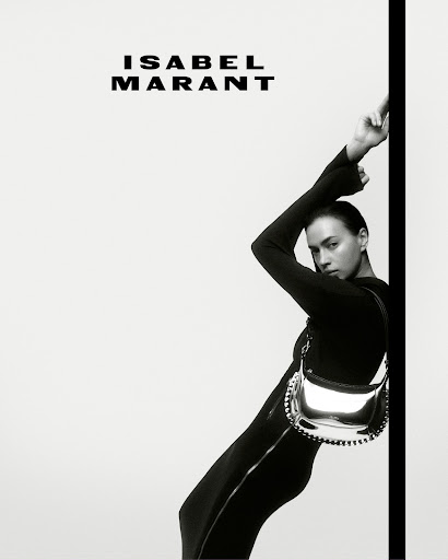 Fashion shot of a woman in a black jumpsuit leaning against a wall with a silver bag, showcasing Isabel Marant branding. High contrast monochrome.