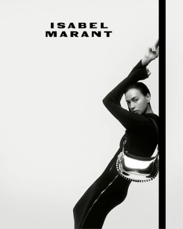 Fashion shot of a woman in a black jumpsuit leaning against a wall with a silver bag, showcasing Isabel Marant branding. High contrast monochrome.