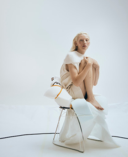 Blonde woman with pale skin sitting with knees drawn up on a wire-frame chair wrapped in white foam by Alice Jacobs