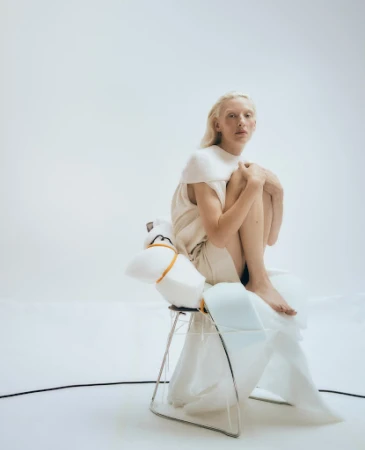 Blonde woman with pale skin sitting with knees drawn up on a wire-frame chair wrapped in white foam by Alice Jacobs