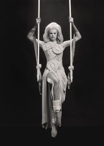 Elegant woman poses on a swing in an elaborately designed dress, complemented by arm coverings, against a stark black backdrop.