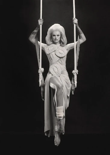 Elegant woman poses on a swing in an elaborately designed dress, complemented by arm coverings, against a stark black backdrop.