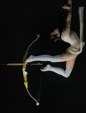 A woman in an aerial pose holds a bow and arrow in a studio against a dark backdrop. The image showcases strength and flexibility.