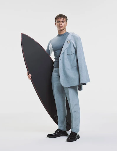 Portrait of a male model in suit jacket and workout shirt holding a surfboard in a studio setting against a white backdrop.
