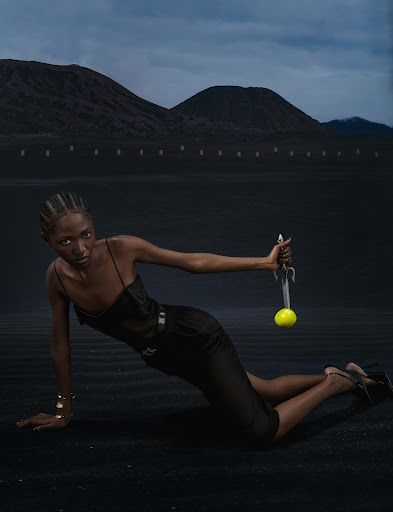 Elegant woman in black outfit holds sword with tennis ball in desert landscape, mountains, and dramatic sky in a fashion editorial.