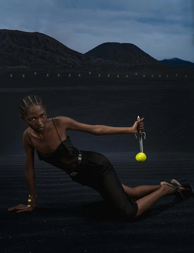 Elegant woman in black outfit holds sword with tennis ball in desert landscape, mountains, and dramatic sky in a fashion editorial.