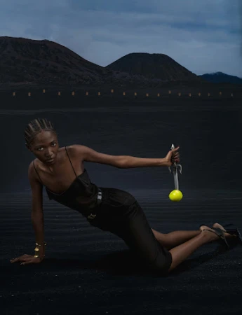 Elegant woman in black outfit holds sword with tennis ball in desert landscape, mountains, and dramatic sky in a fashion editorial.