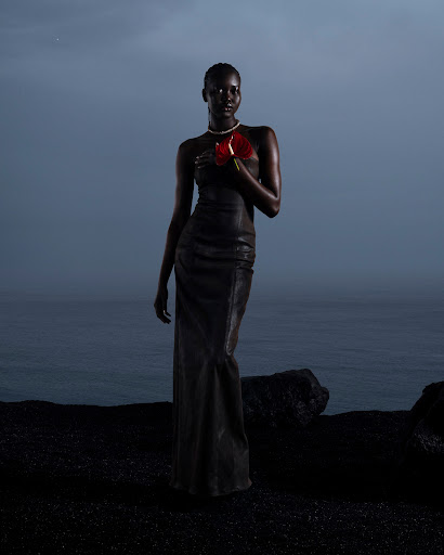 Dark-skinned woman in a black dress holds a red anthurium flower against an ocean and dark sand. She wears a pearl choker.