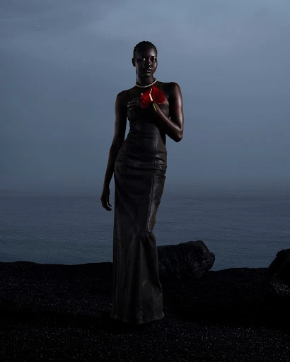 Dark-skinned woman in a black dress holds a red anthurium flower against an ocean and dark sand. She wears a pearl choker.