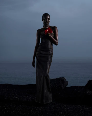 Dark-skinned woman in a black dress holds a red anthurium flower against an ocean and dark sand. She wears a pearl choker.