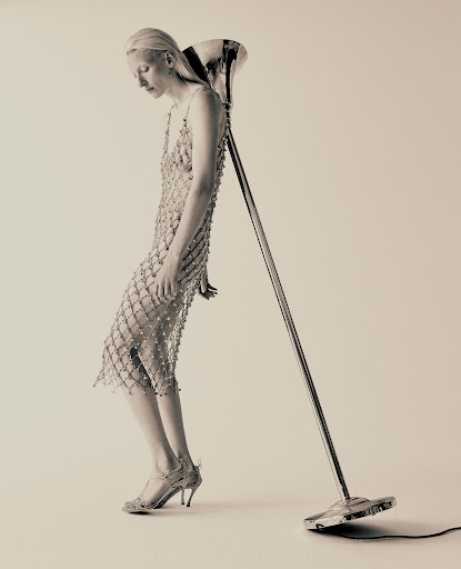 Monochrome fashion shot featuring a fair-skinned woman in a beaded dress leaning against a vintage microphone stand in a light-colored studio.