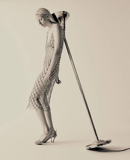 Monochrome fashion shot featuring a fair-skinned woman in a beaded dress leaning against a vintage microphone stand in a light-colored studio.