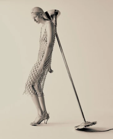 Monochrome fashion shot featuring a fair-skinned woman in a beaded dress leaning against a vintage microphone stand in a light-colored studio.