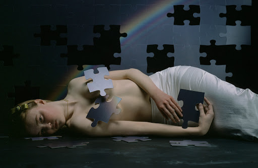Conceptual fine art photograph of a woman laying down surrounded by puzzle pieces against a gray and black puzzle piece background.