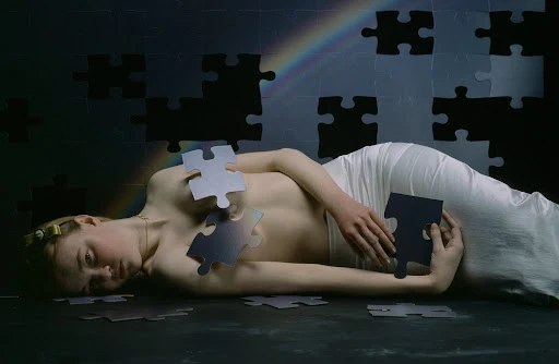 Conceptual fine art photograph of a woman laying down surrounded by puzzle pieces against a gray and black puzzle piece background.