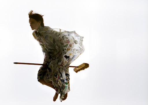Woman in patterned dress appears to fly on a broom against a bright white background in a surreal, fashion-forward, and theatrical image.