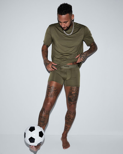 Male model wearing a t-shirt and underwear, posing with a soccer ball against a white background in a studio shot, arms visible, legs tattooed.