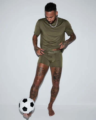 Male model wearing a t-shirt and underwear, posing with a soccer ball against a white background in a studio shot, arms visible, legs tattooed.