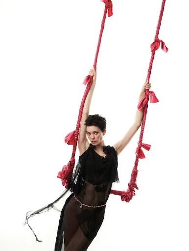 A woman poses on a swing, dressed in a flowing black lace gown against a stark white background in a fashion-forward artistic studio shot.