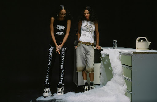 Two young women in graphic print tops and fashionable bottoms pose in a studio kitchen beside white, overflowing foamy suds.