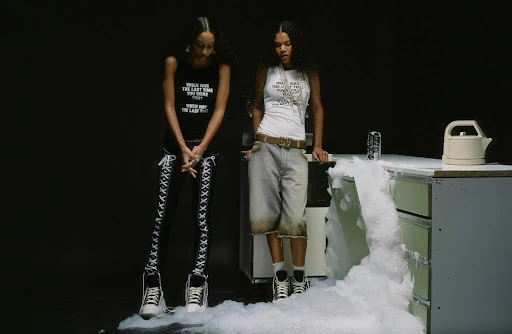 Two young women in graphic print tops and fashionable bottoms pose in a studio kitchen beside white, overflowing foamy suds.