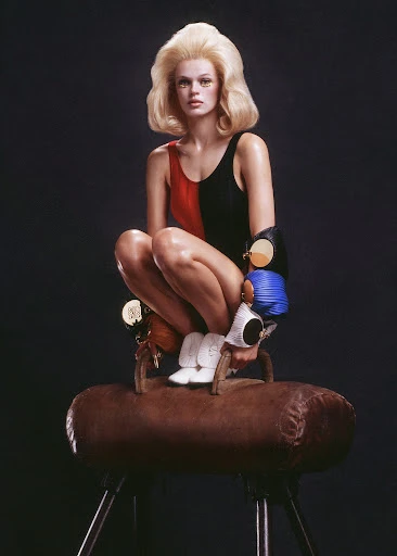 Woman with blond hair in retro swimsuit and sunglasses crouches on gymnastic horse in photo studio with gray backdrop, studio lighting.