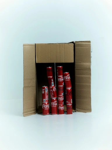 Several red and white Coca-Cola cans are stacked within a cardboard box against a white background, product shot, soda, drink, beverage.
