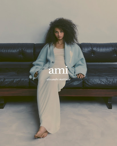 A young adult woman with curly dark hair sitting on a black sofa wearing a light blue cardigan and cream-colored dress in a studio shot.