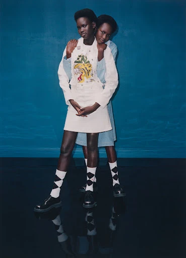 Two black female fashion models in white miniskirts and tops with black and white socks, posing against a dark blue backdrop and reflective floor.