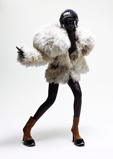 Fashion shot of a Black model in an oversized fur coat and football helmet, posing in heeled boots against a white backdrop.