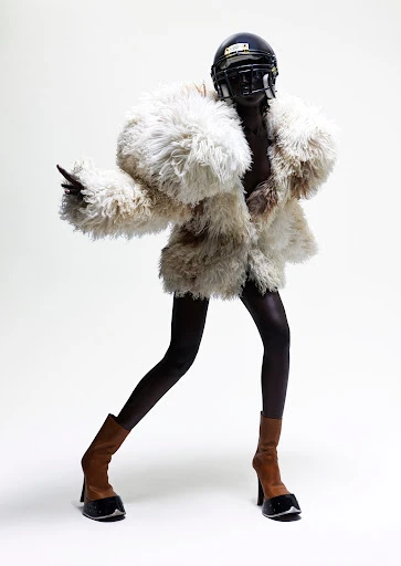 Fashion shot of a Black model in an oversized fur coat and football helmet, posing in heeled boots against a white backdrop.