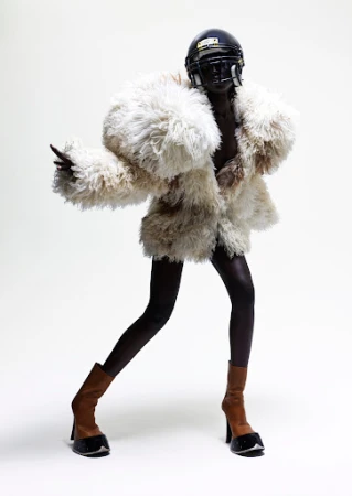 Fashion shot of a Black model in an oversized fur coat and football helmet, posing in heeled boots against a white backdrop.