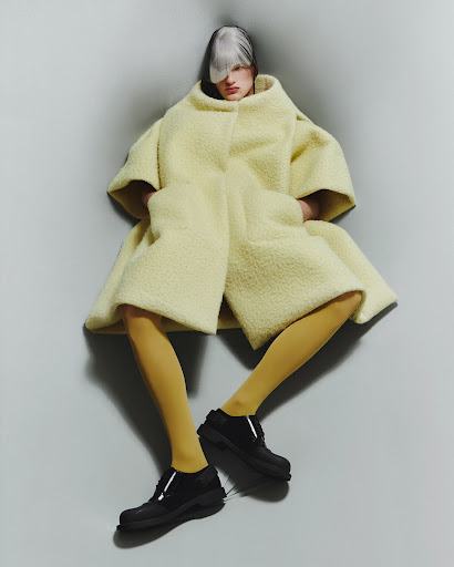 Fashion-forward model posing in a lemon sherpa coat, light yellow leggings, and chunky black shoes against a grey corner backdrop.