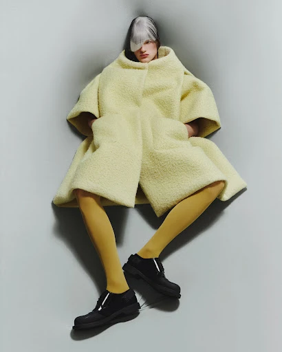 Fashion-forward model posing in a lemon sherpa coat, light yellow leggings, and chunky black shoes against a grey corner backdrop.