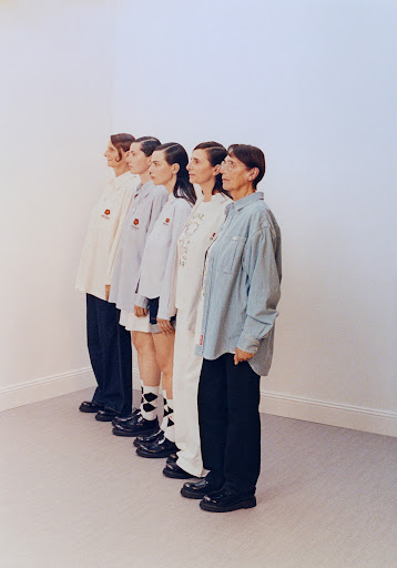 A group of five women stand in a line against a white wall, each wearing different shirts and pants, and black shoes in a photo studio.