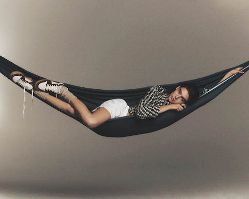 Adult lounging in a hammock, wearing patterned top, white shorts, sneakers and leg warmers, with a neutral background.