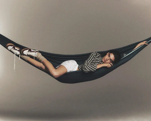 Adult lounging in a hammock, wearing patterned top, white shorts, sneakers and leg warmers, with a neutral background.