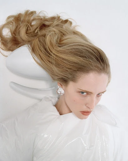 Blonde woman in a white puffy jacket and pearlescent earrings, lying against a white chair with a matching white backdrop.