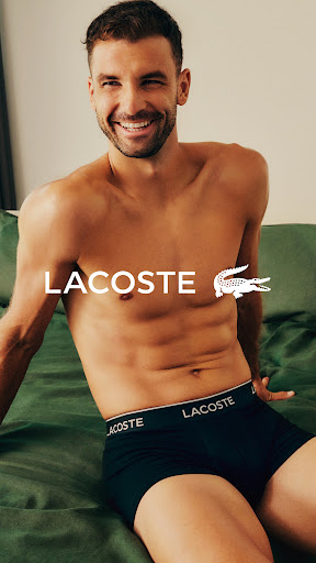 Smiling adult man wearing Lacoste underwear sits indoors in portrait shot, showcasing athletic physique and apparel. Beauty and fashion.