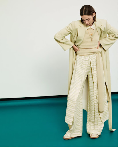 Woman in relaxed fashion outfit with light jacket, ribbed top, and patterned pants in a studio setting against white and green background.