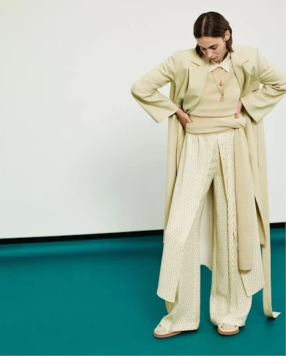 Woman in relaxed fashion outfit with light jacket, ribbed top, and patterned pants in a studio setting against white and green background.