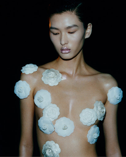 Asian woman with fair skin and dark hair poses with glossy makeup and decorative white camellias against a dark backdrop.