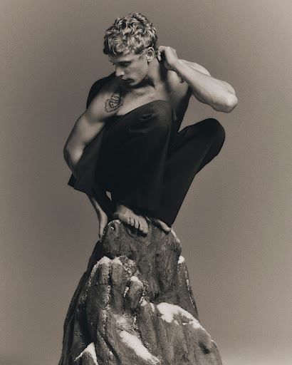 Black and white portrait of a muscular fair-skinned male model squatting on an artificial snow-dusted rock in a photo studio.