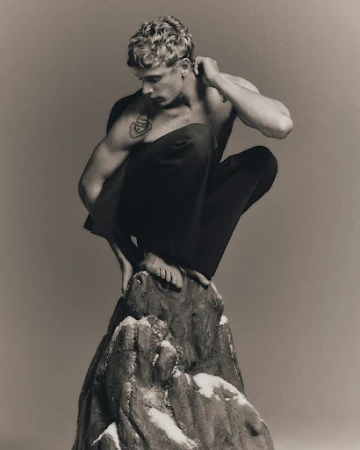 Black and white portrait of a muscular fair-skinned male model squatting on an artificial snow-dusted rock in a photo studio.