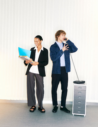 Two business colleagues in a bright office setting; one woman with folders, one man on a landline phone, both in stylish work attire.