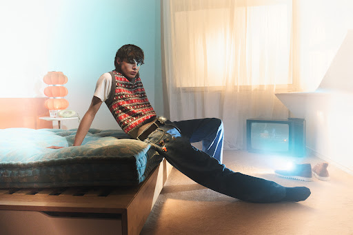 A young man sits on a bed in an indoor setting with a retro TV, wearing jeans and a patterned sweater vest.