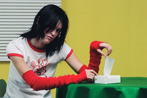 Adult female with dark hair pulls a tissue from a tissue box on a green table, set against a yellow wall and white window blinds.