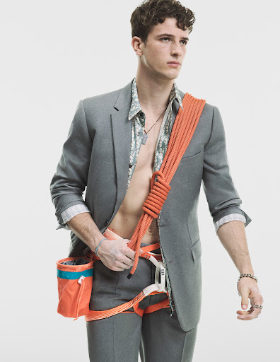 Young man in a suit with climbing gear, including harness, chalk bag, and orange rope slung over his shoulder against a white background.
